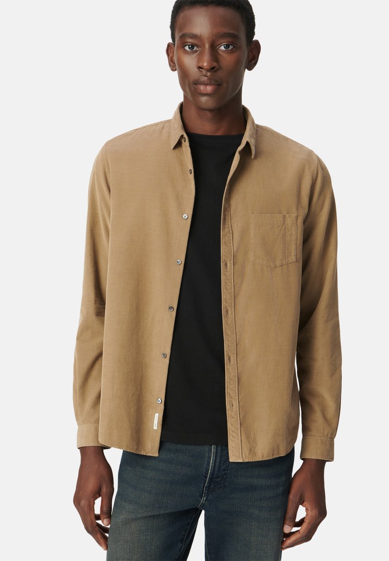 Beżowa koszula z guzikami, z kieszenią na piersi, charakteryzująca się teksturowanym materiałem i zaokrąglonym dołem, noszona na czarnym T-shircie i niebieskich dżinsach.