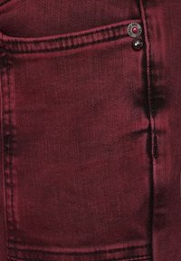 Tejido de denim burdeos con una superficie texturada, que presenta una abertura de bolsillo visible y un botón de metal con detalles de la marca.
