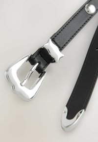 Ceinture en cuir noir avec une boucle en métal argenté brillante et un accent étoile. La ceinture présente une texture lisse et des extrémités pointues avec des embouts métalliques.