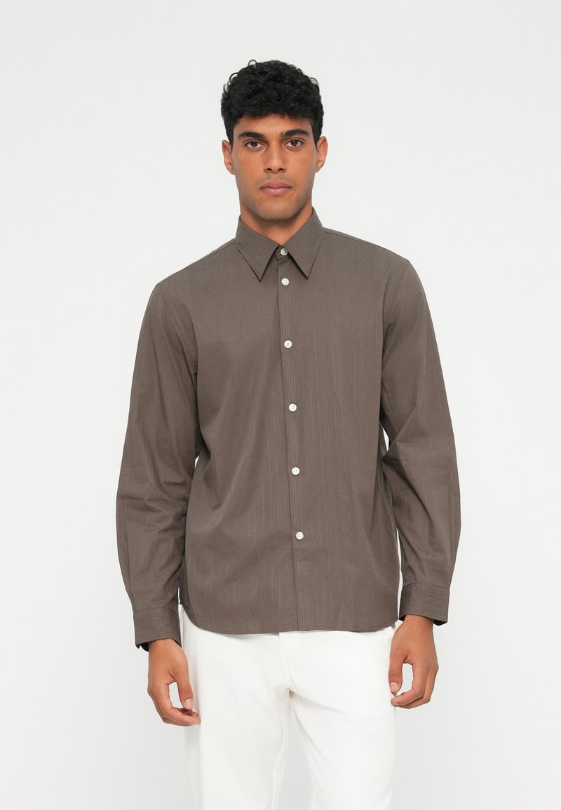 Camisa marrón de manga larga con rayas verticales, cuello clásico y botones blancos. Hecha de una tela suave, combinada con pantalones blancos.