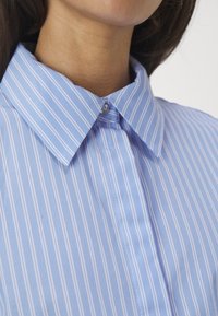 Gros plan sur le cou et une chemise bleue à boutons avec des rayures blanches verticales et un col pointu.