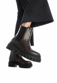 Stivali alla caviglia in pelle marrone con pannello laterale elastico texturizzato, suola nera in gomma spessa e zip laterale. Abbinati a pantaloni neri a sbalzo.