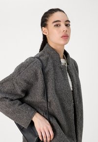 Sivý vlnený kabát s textúrovaným povrchom, so stojatým golierom a prednými cvokmi, doplnený čiernou crossbody taškou.