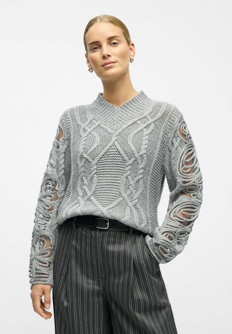 Pull en maille gris clair avec un col en V et des motifs en câble complexes, présentant des manches en transparence. Associé à un pantalon noir à rayures.