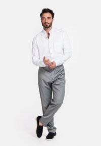 Witte blouse met knoopsluiting en zak, grijsgemaakte pantalon, en zwarte suède instappers. Model staat met één voet naar voren, handen gekruist.