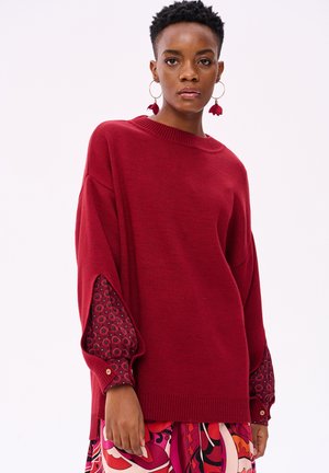 Pull rouge à coupe ample, avec des manches larges arborant une accent floral. Encolure ronde et ourlet côtelé, texture douce.