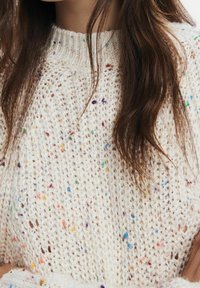 Pull en maille crème avec des flocons multicolores. Présente un col rond côtelé et une coupe ample avec une texture douce et un tissage ouvert.