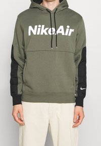 Olivgrön huvtröja med svarta ärmar, prydd med fet vit "NikeAir" text på framsidan och en liten Nike-logotyp på handleden. Tyget är en bomullsblandning.