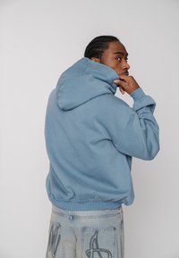 Hellblauer Kapuzenpullover mit lockerem Schnitt, ausgestattet mit gerippten Bündchen an Ärmeln und Saum. Der Stoff wirkt weich und hat eine glatte Textur.