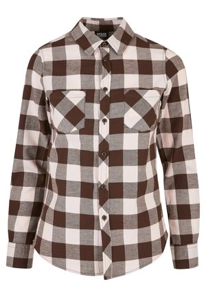 Femme portant une chemise à carreaux marron, blanc et gris boutonnée par-dessus un top marron, manches retroussées et pantalon noir.