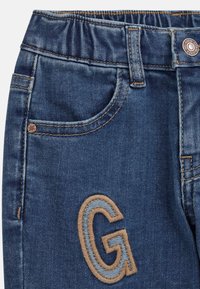 Denim farkut joustavalla vyötäröllä, tummansiniset, joissa on etutasku ja vaalean ruskea brodeerattu "G"-logo.