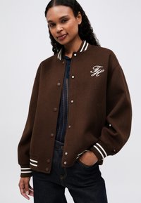 Mujer con una chaqueta de varsity marrón con rayas blancas en el cuello y los puños, bordada con "TH" en el pecho, top azul oscuro y jeans oscuros.