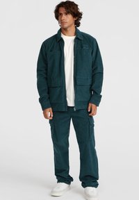 Teal corduroy jack met twee borstzakken, gecombineerd met bijpassende cargo-broek. De outfit heeft een relaxte pasvorm en een wit t-shirt eronder.