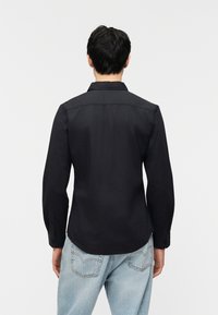 Άνδρας με κοντά σκούρα μαλλιά, φορώντας μαύρο πουκάμισο με μακριά μανίκια και ανοιχτό μπλε τζιν, στέκεται με την πλάτη προς το μέρος του θεατή πάνω σε λευκό απλό φόντο.