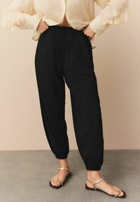Sorte, lette, elastiske bukser med en tapered ben-design. Båret med en cremefarvet bluse og guldstribbede sandaler, mod en neutral baggrund.