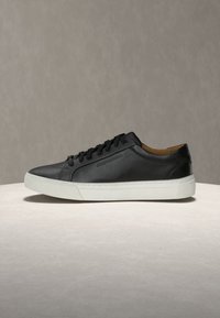 Μαύρο δερμάτινο sneaker χαμηλού προφίλ με μαύρα κορδόνια και λευκή καουτσούκ σόλα σε ουδέτερη επιφάνεια και φόντο.