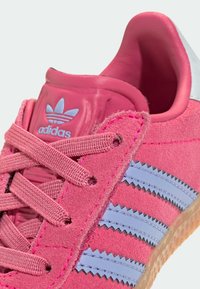 Sapatilha de camurça rosa da Adidas com riscas azuis, atacadores texturizados e um colarinho acolchoado. Sola de borracha com um acabamento de borracha.