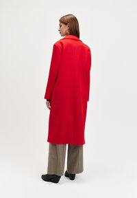 Długi czerwony płaszcz z strukturalnego materiału, z klapami, długimi rękawami oraz prostym krojem, zestawiony z szerokimi spodniami w kratę.