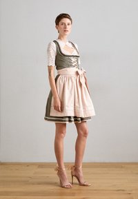 Tradicionalna dirndl obleka v sivi barvi s svetlo rožnato predpasnikom; ima čipkaste rokave, prilagojen korzet in naglavne trakove. Visoke sandale dopolnjujejo videz.