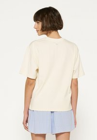 Lyse beige bomulds t-shirt med rund halsudskæring, korte ærmer og løstsiddende pasform. Båret med blå og hvide stribede shorts.