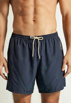 Marineblaue Badeshorts mit grüner Kaktus-Stickerei, weißem Kordelzug im Bund und seitlicher Reißverschlusstasche, getragen von einem oberkörperfreien männlichen Model.