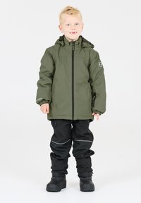 Giacca impermeabile verde con zip frontale e cappuccio, indossata sopra pantaloni neri. Presenta polsini elasticizzati, dettagli riflettenti e robusti stivali neri.