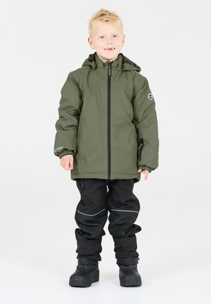 Giacca impermeabile verde con zip frontale e cappuccio, indossata sopra pantaloni neri. Presenta polsini elasticizzati, dettagli riflettenti e robusti stivali neri.
