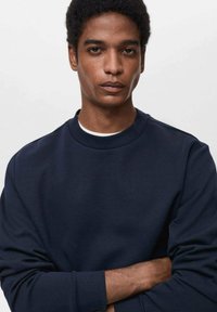 Temno modra senca pulover iz mehke tkanine, z okroglim izrezom in dolgimi rokavi. Minimalistična zasnova, gladka tekstura in brez vidnih vzorcev.