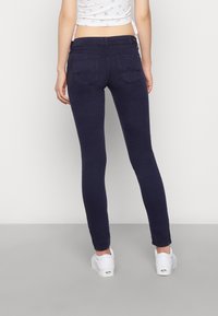 Jeans skinny en color navy con una textura suave y bolsillos traseros, combinados con zapatillas blancas. Cintura plana y diseño ajustado en los tobillos.