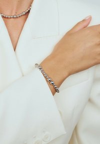 Silverfärgade pärlhalsband och matchande armband bärs på en persons handled och hals över en vit blazerärm.