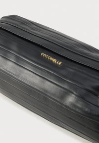 Μαύρο δερμάτινο τσαντάκι χειρός με υφή και χρυσλό λογότυπο Coccinelle σε λευκό φόντο.