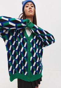 Pletený kardigan s geometrickým vzorem v zelené, modré, černé a bílé barvě. Disponuje výstřihem do V, zapínáním na knoflíky a žebrovanými manžetami a spodním lemem.