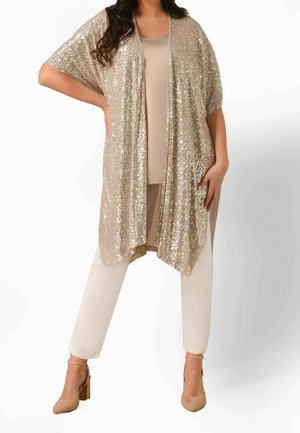 Vrouw draagt een beige top en broek met een glanzende, losse, goudkleurige cardigan met pailletten en nude pumps.
