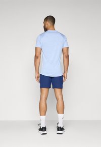 Un t-shirt d'athlétisme à manches courtes bleu clair avec des manches raglan, associé à un short navy. Le mannequin porte des chaussettes blanches et des chaussures de course noires.