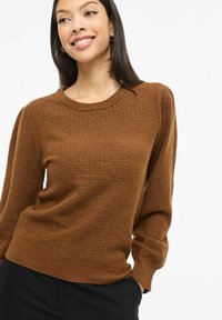 Brun strikket genser med teksturert mønster, rund hals og lange ermer. Stylet over svarte bukser med en figursydd kant.