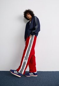 Navy bomber jacket with three gray stripes on sleeves, red track pants with black and white side stripes and snaps, blue sneakers with three stripes.