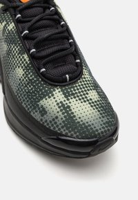 Schwarzer und grüner Sneaker mit einem strukturierten Obermaterial und einem Punktmuster, schwarzen Schnürsenkeln und einer glänzenden schwarzen Sohle. Akzentuiert durch ein orangefarbenes Detail.