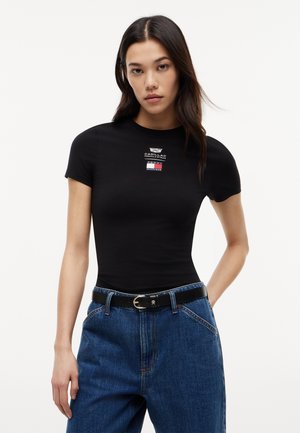 Mujer con cabello largo y oscuro, vistiendo una camiseta negra ajustada con pequeños logotipos de Cadillac y Tommy Hilfiger, jeans azules y un cinturón negro.