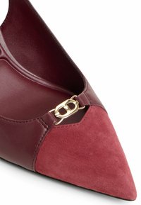 Sko med spetsig tå som har ovandel i burgundy läder och mocka, med en guldfärgad metallaccent vid den främre utskärningen. Slät innerfoder.