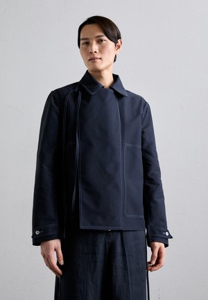 Veste marine en tissu lisse, avec des coutures contrastantes bleu clair, un col et deux poches avant. Manches longues avec poignets boutonnés.
