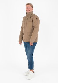 Giovane in piedi, indossa una giacca beige, jeans blu e sneakers bianche, su uno sfondo bianco semplice.