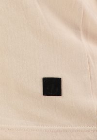 Tessuto rosa chiaro con una texture a righe, dotato di un'etichetta quadrata nera in rilievo con il nome del marchio e un simbolo nell'angolo in basso a destra.
