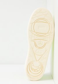 Semelle d'une chaussure de couleur claire présentant des motifs géométriques en relief, comprenant des cercles, une étoile et des lignes croisées, sur un fond blanc.