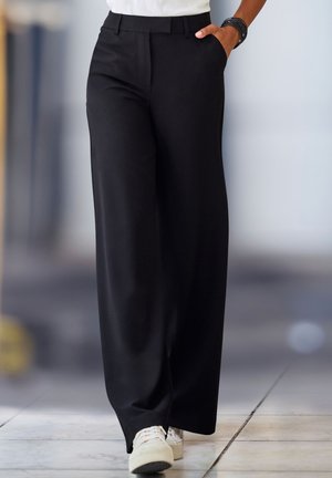 Person trägt schwarze, weit geschnittene Hose, weiße Turnschuhe und ein weißes Oberteil mit einem schwarzen Armband, geht auf gefliestem Boden.