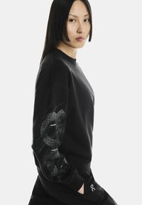 Fekete pulóver kerek nyakkal, textúrált grafikai mintával és logóval a hajtón. Az anyag lágy és kényelmes hatását kelti.