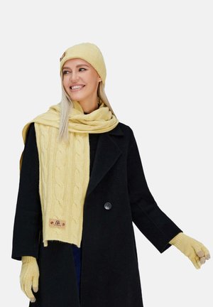 Berretto giallo lavorato a maglia, sciarpa con motivo a treccia e guanti, abbinati a un cappotto nero. Dettagli degni di nota includono un'etichetta sulla sciarpa.
