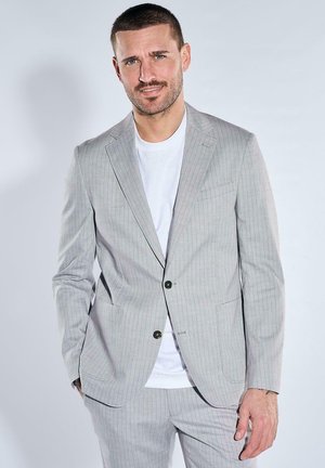 Mann trägt hellgraue Nadelstreifen-Anzugjacke und passende Hose über einem weißen T-Shirt, steht vor einfarbigem Hintergrund.