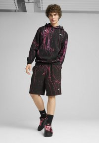 Sudadera negra con capucha y pantalones cortos con un patrón de manchas rosa, que cuenta con un bolsillo frontal y detalles de logo. Combinado con zapatos y calcetines negros.