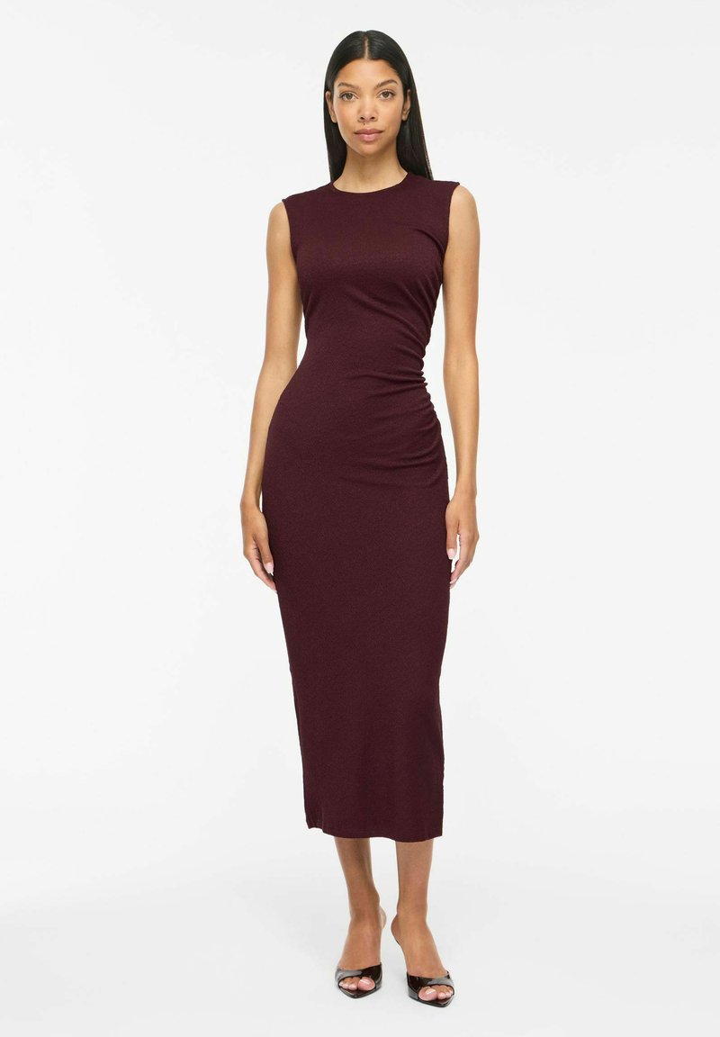 Femme portant une robe midi sans manches, ajustée, bordeaux foncé, et des talons noirs ouverts à l'avant, debout sur un fond blanc uni.