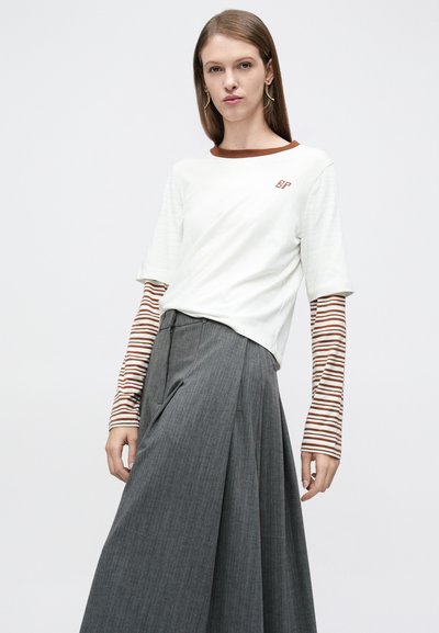 Λευκό μπλουζάκι με καφέ λεπτομέρειες και λογότυπο, φορεμένο πάνω από μακρυά ριγέ μανίκια. Μια γκρι πλισέ φούστα ολοκληρώνει το σύνολο.